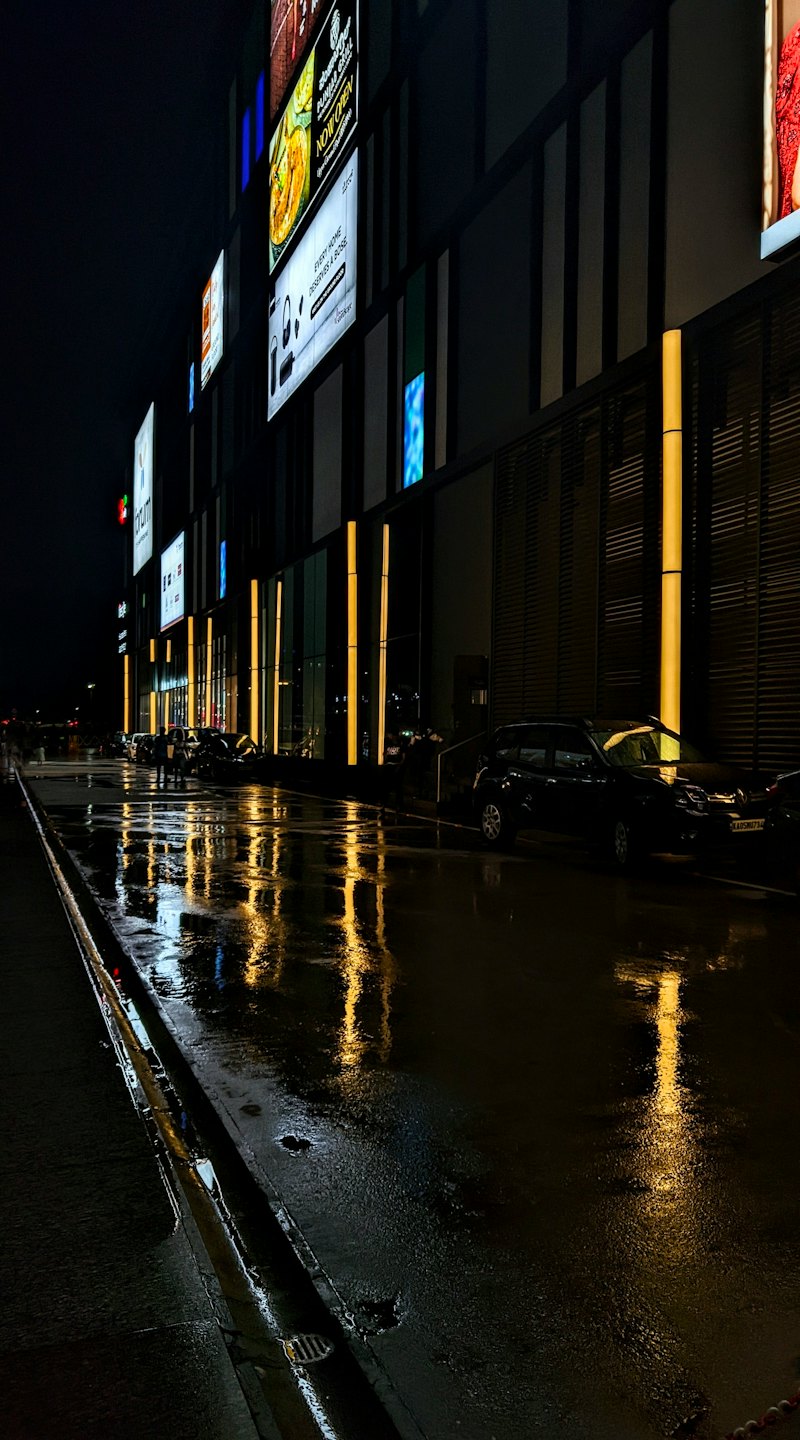 Le luci di un moderno edificio commerciale si riflettono su una strada cittadina bagnata dalla pioggia.
